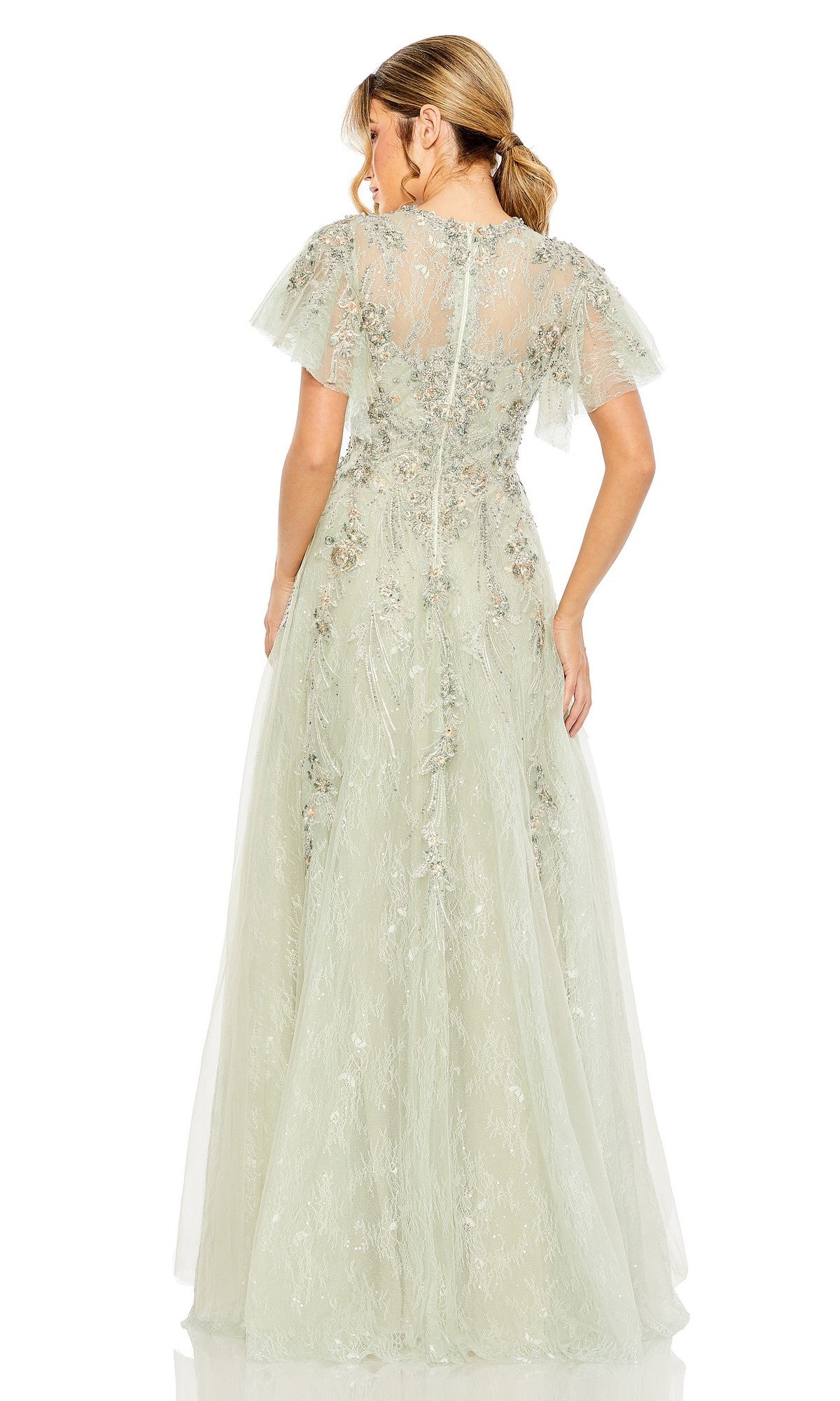 A woman faces away, wearing the Mac Duggal 20450—a light green, floor-length formal dress featuring floral embroidery and sheer short sleeves. Her hair is styled in a low ponytail.